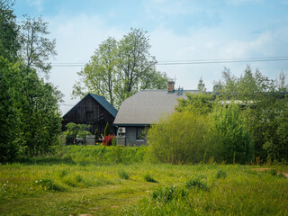 Obraz premium A farmhouse with a modern roof stands next to a weather-beaten wooden barn, surrounded by green grass, trees, and spring foliage under a blue sky.