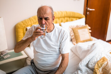 Senior man drinking water in the morning while sitting on bed in his cozy bedroom. 