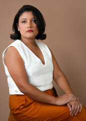 Elegant businesswoman in white top and orange pants sitting and looking at camera. Ideal for professional development, career, personal growth. Copy space on mocha mousse color background.