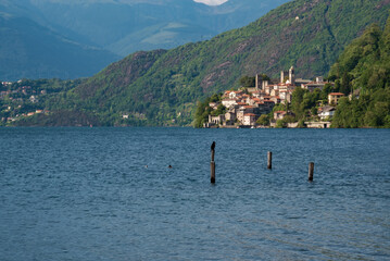 A scenic lakeside village nestled at the foot of lush green hills, with majestic mountains rising in the background under a partly cloudy sky