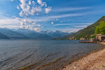A scenic lakeside village nestled at the foot of lush green hills, with majestic mountains rising in the background under a partly cloudy sky