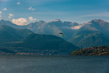 A scenic lakeside village nestled at the foot of lush green hills, with majestic mountains rising in the background under a partly cloudy sky
