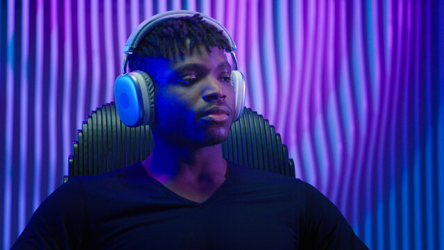 Portrait calm dreaming relaxing tranquil peaceful resting African American man guy male user futuristic indoor futurism listening music in modern headphones listen audio song earphones sound dark room