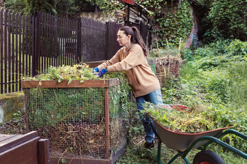 Woman adding garden waste and weeds to compost bin in backyard, sustainable gardening and organic...