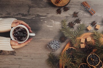 Christmas flat lay. Female hands in a white sweater with coffee cup and frosty lettering in it. Spruce twigs, anise, brown sugar, round cutting board on a wooden table.