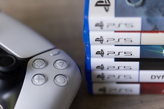 Brecht, Belgium - 18 april 2025: A stack of sony playstation 5 videogame cases or boxes lying on a wooden table next to a wireless PS5 controller. The pile of PS5 games are of multiple genres.