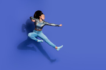 Energetic young woman in casual stylish outfit jumping against a vibrant blue background,...