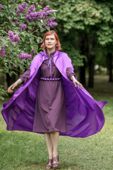 A beautiful young woman with red hair, against a background of blooming lilacs, in a cloak and hood. Medieval fantasy costume. Fairy tale heroine, princess, duchess.