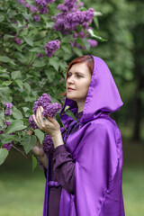 A beautiful young woman with red hair, against a background of blooming lilacs, in a cloak and hood. Medieval fantasy costume. Fairy tale heroine, princess, duchess.