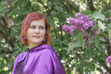 A beautiful young woman with red hair, against a background of blooming lilacs, in a cloak and hood. Medieval fantasy costume. Fairy tale heroine, princess, duchess.