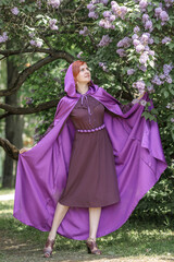 A beautiful young woman with red hair, against a background of blooming lilacs, in a cloak and hood. Medieval fantasy costume. Fairy tale heroine, princess, duchess.