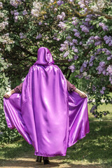 A beautiful young woman with red hair, against a background of blooming lilacs, in a cloak and hood. Medieval fantasy costume. Fairy tale heroine, princess, duchess.