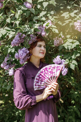 A beautiful young woman with red hair, against a background of blooming lilacs, in a cloak and hood. Medieval fantasy costume. Fairy tale heroine, princess, duchess.