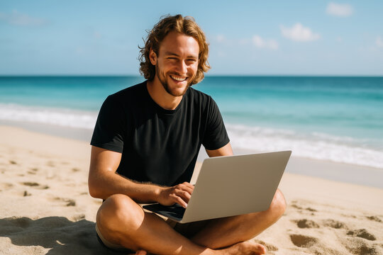 Smiling young man sitting cross legged on sandy beach using laptop while working remotely near turquoise sea - Powered by Adobe