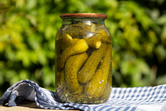 Jar of homemade preserves cucumbers on a wooden table against a background of greenery.