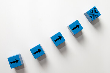 Blue wooden blocks with arrows forming an ascending path towards a target icon.Concept of steps to success,business strategy,achieving goals,progress,growth,and development.White background, flat lay.