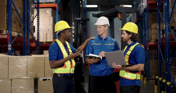 Three male warehouse workers from diverse ethnicities actively planning teamwork using laptop and clipboard while standing beside forklift during logistics operation inside large industrial warehouse - Powered by Adobe