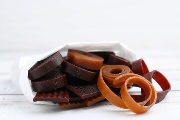 Tasty fruit leather rolls in paper bag on white wooden table, closeup