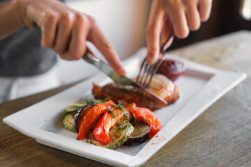 Girl cuts a cooked pork steak served on a white plate with grilled vegetables. Young woman using knife and fork to cutting meat eating in a restaurant. Balanced diet, cooking, culinary. Food concept