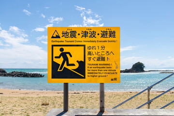 Large notice board on a beach in Japan warning for Tsunamis in case of an earthquake with sea, rock formation in shape of a wave and horizon in background (Japanese text translates on warning sign)