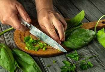 Hands are skillfully cutting fresh greens and herbs on a wooden board, with vibrant leaves and chopped vegetables arranged nearby, showcasing culinary preparation