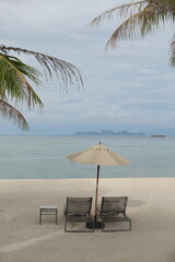 Tropical beach scene with two lounge chairs and a beige umbrella facing the calm ocean. Framed by palm leaves, the tranquil setting features a distant island and a sailboat under cloudy skies.