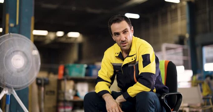 Caucasian male warehouse worker sitting in chair resting and cooling down while holding hardhat after long shift in industrial storage facility during break period inside distribution center