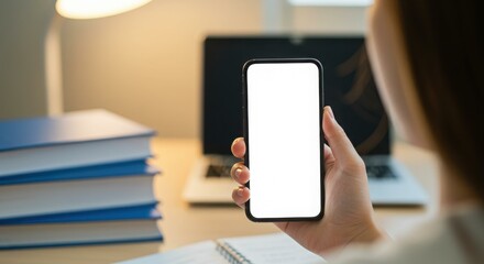 Woman holds blank smartphone screen with laptop and books in warm, inviting study space