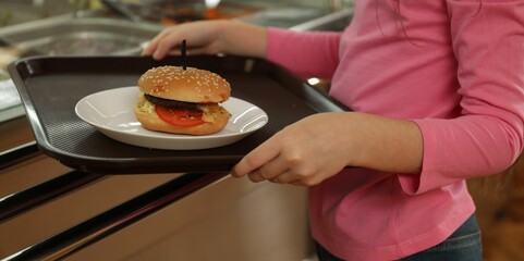Little girl with plastic tray and burger near serving line in canteen, closeup. School food