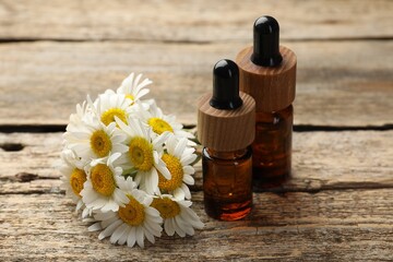 Bottles of essential oil and chamomile flowers on wooden table, closeup