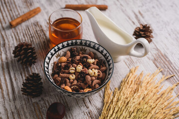 Bowl of healthy breakfast cereal with nuts, dried fruit, milk, and honey on a rustic wooden table, surrounded by natural elements. Cozy morning.