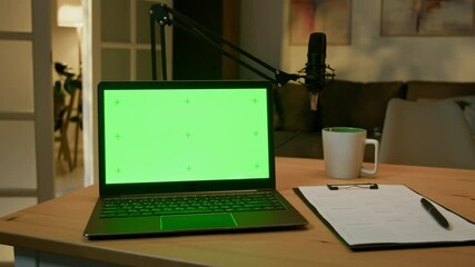 Black laptop with green screen and clipboard with script on table, professional mic set in background