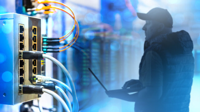 Engineer with laptop in a server room monitors network switch connections. Focus on Ethernet and fiber optic cables. Activity: network diagnostics, IT infrastructure.