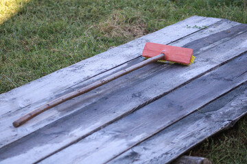 A broom with a red and yellow brush head placed on a wooden surface, with grass visible in the background. The contrast between the structured object and natural elements adds visual interest	
