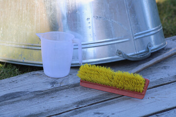 A plastic measuring cup and a yellow brush with a red handle placed on a wooden surface, with a metallic container in the background. The objects suggest practical use in a workshop or household setti