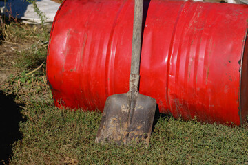 A red barrel with a shovel leaning against it, placed outdoors. The worn metal and utilitarian tools suggest practical, hands-on work	
