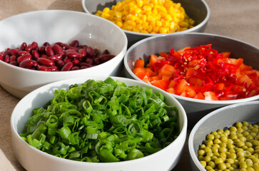 Close-up of several white and grey bowls filled with fresh, colorfully sliced ​​green onions, red beans, diced red peppers, yellow sweet corn and green peas ready for cooking.