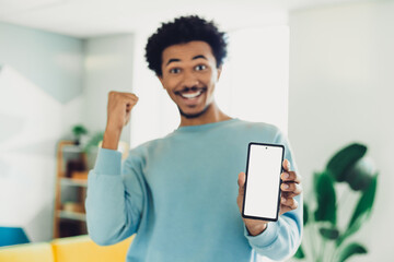 Young smiling man indoors happily displaying smartphone in a bright casual setting with modern home...
