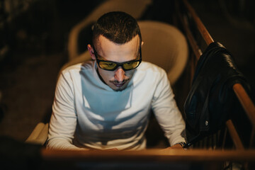 A man with short hair and tinted glasses is seated indoors, focused on his laptop. The setting is...