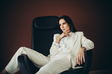 Confident businesswoman in a formal white suit sitting thoughtfully on a chair with a brown...