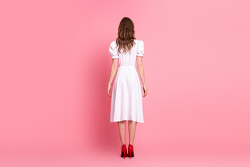 Young woman in a stylish white dress and red heels standing against a pink background showcasing...