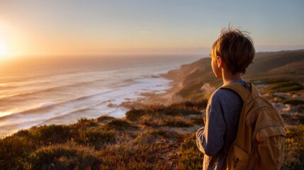 A young boy stands on a coastal cliff, looking out at the waves crashing against the shore. He carries a backpack as the sun sets, casting golden hues over the ocean and landscape