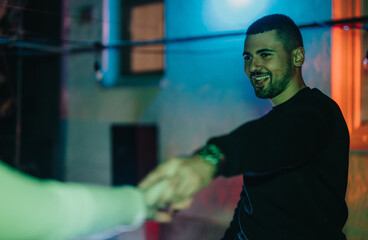 The image shows a smiling young man extending his hand, surrounded by vibrant colorful lighting, evoking feelings of camaraderie and joy in a lively social gathering.