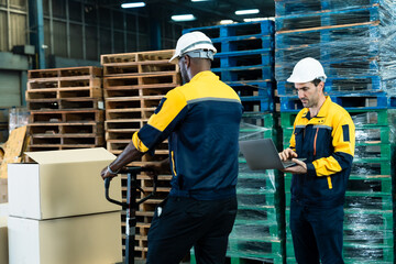 African adult male operating hand pallet jack moving cardboard boxes while caucasian adult male checking data on laptop in warehouse showing coordination and productivity during shipment process