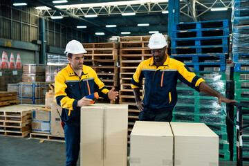 Caucasian adult male worker explaining packaging method to african adult male while standing next to cardboard boxes in warehouse, working together as logistics team in industrial cargo zone