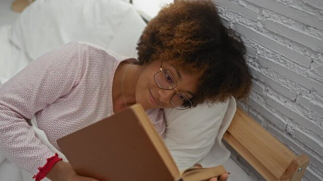 Woman reading book in cozy bedroom with curly hair and glasses, lying on bed with white pillows against a white brick wall, capturing a peaceful indoor moment at home. - Powered by Adobe