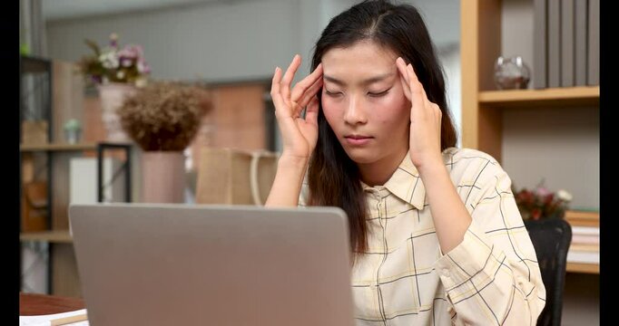 Young Asian adult woman with long dark hair sitting at office desk touching temples with both hands, eyes closed with stressed expression, laptop in foreground, conveying fatigue or frustration 