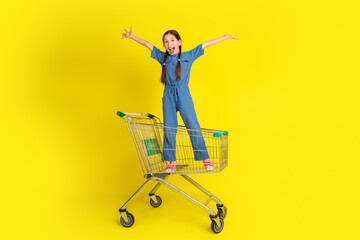 Happy young girl in denim overalls stands stylishly in a shopping cart against a vibrant yellow...