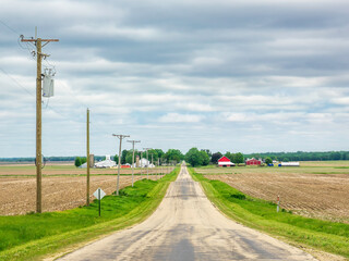Perspective of a rural road with farmland and farm buildings on either side on an overcast afternoon in May, north central Illinois, for motifs of the American Heartland