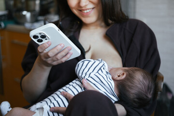 Beautiful young woman is breastfeeding her baby boy at home, close-up view. She is working online at the same time, remote work from home. Chatting with friend, having online shopping. 